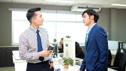 Two male colleagues employees cooperating in office talking working together at workplace, smiling and drinking coffee