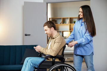 A cute young man in a wheelchair is holding a mobile phone while his girlfriend helps him move around the apartment