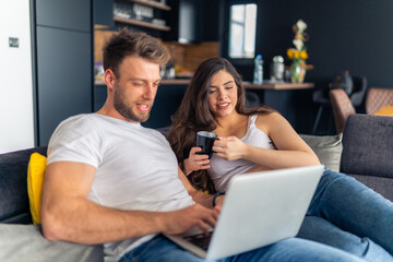 Expecting new parents are sitting on the couch planning the decor for the new children's room, the laptop is on his lap while she holds a cup of tea in her hand