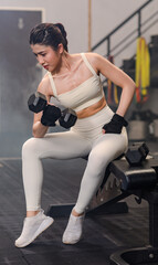 Fototapeta na wymiar Asian young female fit strong body sporty athletic fitness model in sport bra legging gloves sitting posing holding lifting metal dumbbell weight training arms bicep exercise at CrossFit gym.
