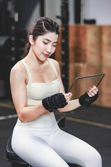 Asian young muscular fit strong body sporty athletic sexy female fitness model sitting smiling taking break holding looking at tablet computer mockup after workout exercising weight training in gym