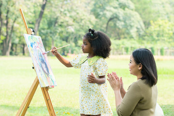 Cute African daughter painting coloring watercolor on canvas in green park on summer. Asian mother...