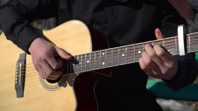 A young man plays the acoustic guitar while sitting in the park. A man plays the guitar close-up. The camera is moving.