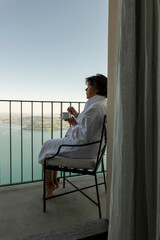 Woman with Bathrobe Sitting and Drinking a Coffee and Enjoy the View From Balcony with Mountain and Lake in a Sunny Summer Day on Lake Lucerne From Burgenstock, Nidwalden, Switzerland.