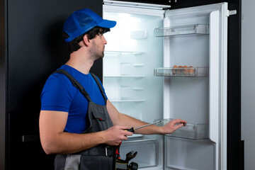 Male technician in uniform repairs a broken refrigerator in the kitchen. 