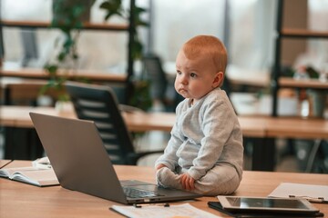 Sitting on the table by laptop. Cute little baby boy is in the modern office