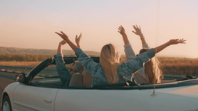 Young people in a white car with open top drive at sunset along a country road and raise their hands in the air in pleasure. Group of young people enjoys a vacation in a car country trip