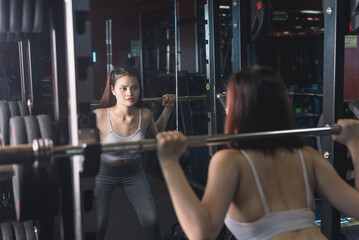 A young and fit woman does barbell squats in front of the mirror at the gym. Using the bar weight only.