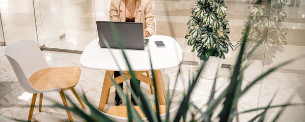 A business woman sits in a cafe and works at a computer. She is wearing a beige jacket.