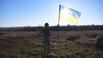 Soldier of ukrainian army going with lifted banner on the field at sunny day. Male military in uniform walks with waving flag of Ukraine at meadow. Victory against russian aggression. Slow motion