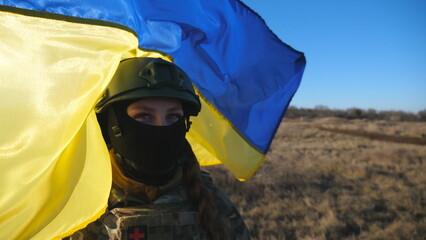 Female military medic of ukrainian army looks into camera against waving blue-yellow banner. Portrait of girl in camouflage uniform and helmet holding flag of Ukraine at field on sunny day. Dolly shot