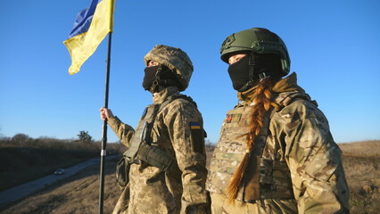 Young soldiers of ukrainian army standing at peak of hill with raised flag of Ukraine. Military couple in camouflage uniform taking each other hands as a symbol of support. Victory at war. Crane shot