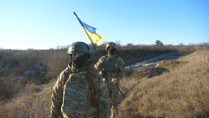 Young woman and man in military uniform going with national banner at meadow. Female and male soldier of ukrainian army walking with flag of Ukraine at field. Victory against russian aggression