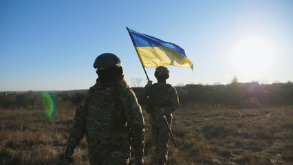Young woman and man in military uniform going with national banner at meadow. Female and male soldier of ukrainian army walking with flag of Ukraine at field. Victory against russian aggression - obrazy, fototapety, plakaty