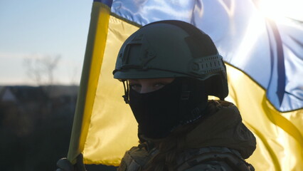 Female military of ukrainian army looks into camera against waving blue-yellow banner. Portrait of girl in military helmet and balaclava with national flag on sunny day. Russian invasion of Ukraine