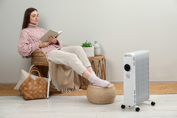 Woman reading book near modern portable electric heater indoors