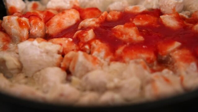 Frying chopped chicken breast pieces pouring with red sauce. Close-up. Roasting chicken fillet in black frying pan. Stewed poultry Recipe. Fresh raw white meat cooked in a steamer. Diet food. Overhead