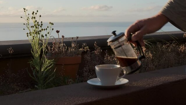 Caf&eacute; caliente humeante de cafetera de &eacute;mbolo sobre el muro de una jardinera con luz de amanecer en una terraza con vistas al mar y mano que coge la taza