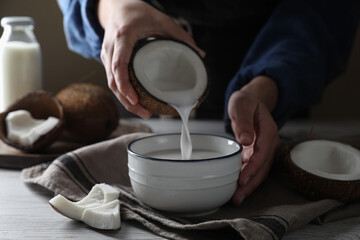 Woman pouring coconut milk into bowl at white wooden table, closeup