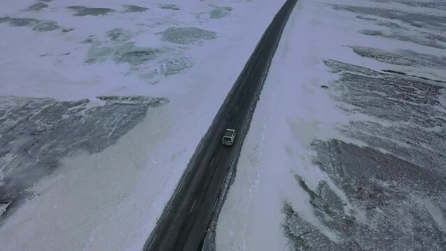 Voiture van suivi par un drone sur une route enneigé au milieu d'une nature sauvage d'Islande recouverte par la neige avec une route noire et des fleuves et lac  gelés 