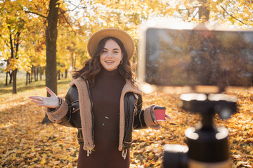 Female blogger recording video outdoors. Beautiful young girl smiling and looking at the camera standing in the autumn park, talking and gesticulating with both hands. Blogging, video blog