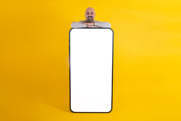 Big smartphone mock up, caucasian bald handsome man leaning big smartphone mock up. Yellow studio background copy space. App ad concept idea. White screen template. Giant cell phone for advertising.