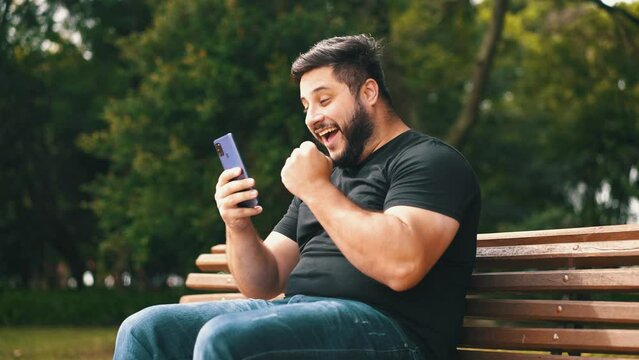 Young man celebrating good news recieved on his cell phone while seated at a park bench