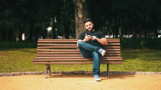 Young mand using his cell phone smiling seated alone on wood park bench on sunny day