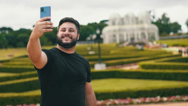 Young man with smiling and taking a selfie at a beautiful park botanical garden with a big greenhouse on the background