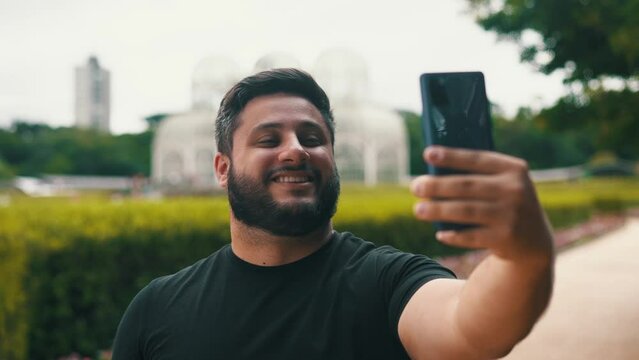 Young bearded man smiling and taking a selfie at a botanical garden with a big greenhouse on the background located in Curitiba, Brazil