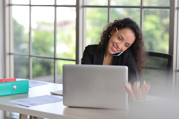 Attractive professional latin female employee worker sitting, using laptop computer with paperwork at home workplace. Businesswoman using speaker phone on smart mobile phone while working.