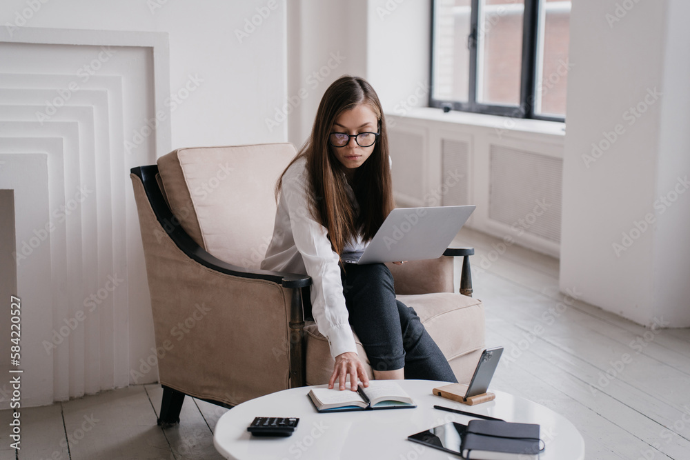 Wall mural Busy brunette American young businesswoman sitting in armchair holds laptop opens diary, remote works home. Pensive student girl in glasses distant learning. Purposeful manager overloaded.