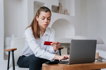 Perplexed young businesswoman in white shirt and black pants using laptop holds credit card makes online purchase. Debt, payment. Hispanic female entrepreneur working at office. fatigue concept.