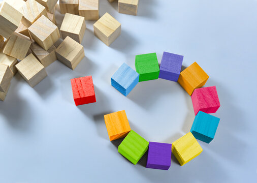 Cubes in different colors together form a circle with a gap, one is red and stands separate, heap of natural wooden cubes in the corner, light blue background, copy space