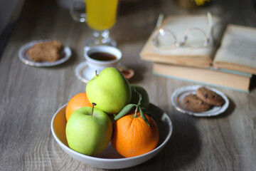 Cup of tea, plates with cookies, glass of orange juice, books, reading glasses, bowl of fruit and candles on the table. Selective focus.