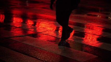 Person walks slowly on wet road crosswalk with red light reflecting on ground
