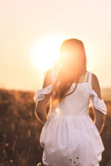 Fototapeta na wymiar Rear view of young woman in summer white dress walking on sunset background. Attractive girl looking into the distance at nature