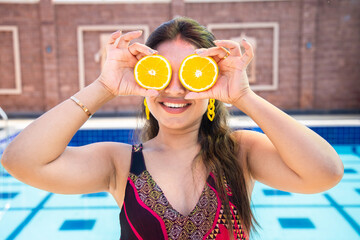 Beautiful happy young indian woman put two pieces of orange slices on her eyes as eye wear having fun with tropical juicy fruit in hot sunny day. Summer food concept.