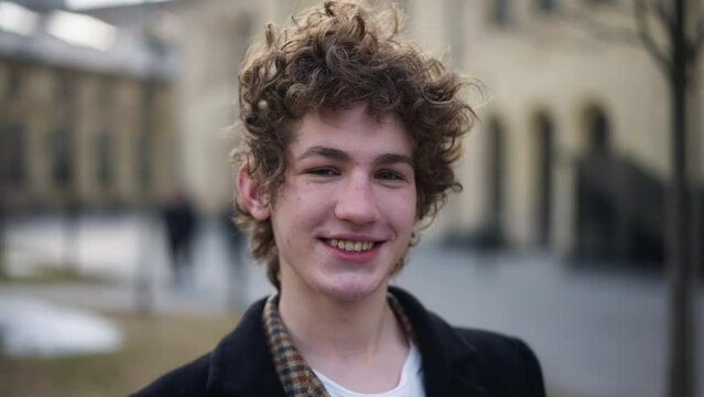 Close-up face of smiling happy man touching curly hair laughing looking at camera. Front view headshot of confident cheerful Caucasian young guy posing outdoors on city street having fun