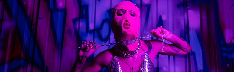 passionate woman in balaclava posing with silver chain near graffiti in purple light, banner.