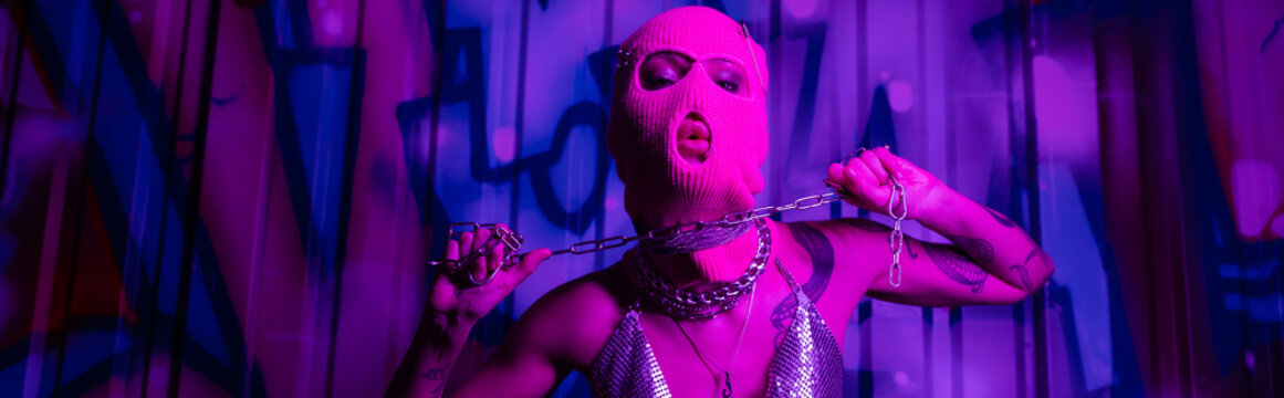 passionate woman in balaclava posing with silver chain near graffiti in purple light, banner.