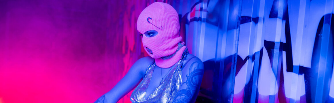 anonymous tattooed woman in balaclava near wall with graffiti in blue and pink lighting with smoke, banner.
