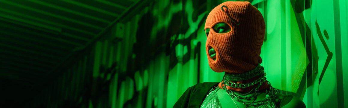 sexy anonymous woman in orange balaclava and silver necklaces looking away in green light near wall with graffiti, banner.