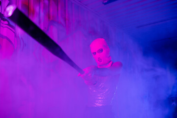 provocative woman in balaclava holding baseball bat in outstretched hands while standing in purple fog.