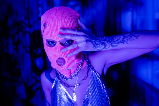 provocative woman in balaclava and metallic top with necklaces posing with hand near face in blue and purple lighting.