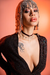 portrait of sensual woman with sexy tattooed body posing in black dress and silver wig with rhinestones on coral pink background.