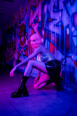 full length of passionate woman in balaclava and leather skirt with boots posing near wall with graffiti in blue and purple light.