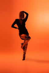 full length of sexy model in fashionable boots and black dress posing on one leg on orange background.
