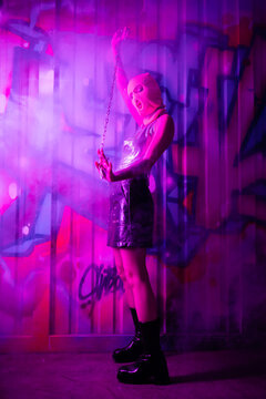 full length of woman in balaclava and leather boots posing with chain near colorful graffiti in purple light with smoke.