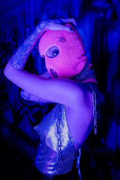 provocative tattooed woman in metallic top and knitted balaclava looking at camera in blue and purple neon light.